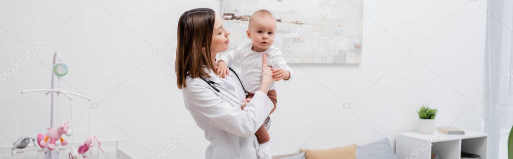Young family doctor in white cot holding baby boy at home, banner 