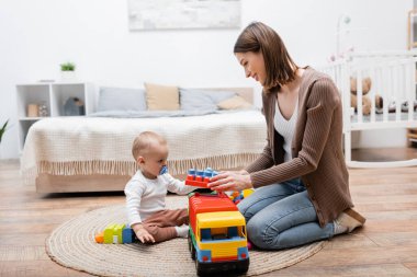 Emzik taşıyan bebek annesinin yanında oturuyor ve elinde yapı taşları var. 
