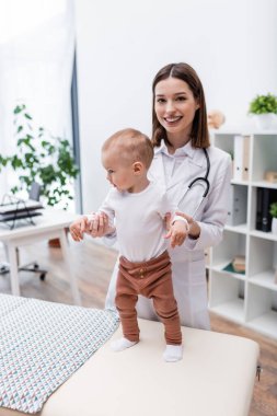 Gülümseyen aile doktoru hastanedeki kanepede bebeğin elini tutuyor. 