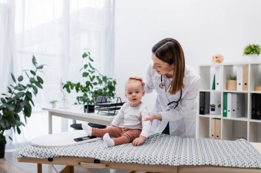 Gülümseyen aile doktoru klinikteki kanepede bebeğin kafasına dokunuyor. 