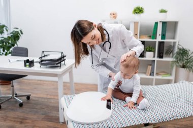 Klinikteki tıbbi kanepede bebeği elektronik ölçekte muayene eden olumlu doktor.