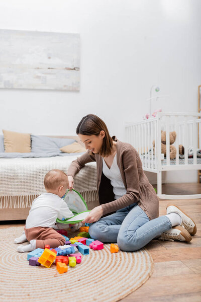 Smiling mom holding bag with building blocks near baby son in bedroom 