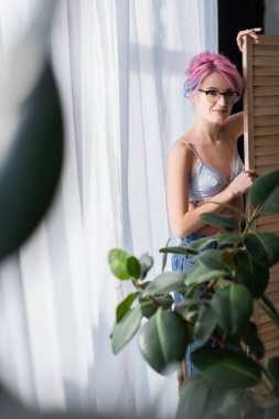 Gülümseyen, boyalı saçlı genç bir kadın katlanan perdenin yanında duruyor.