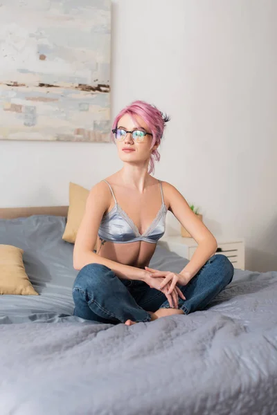 ピンク色の髪をした若い女性がサテンブラジャーとジーンズにベッドで座り — ストック写真