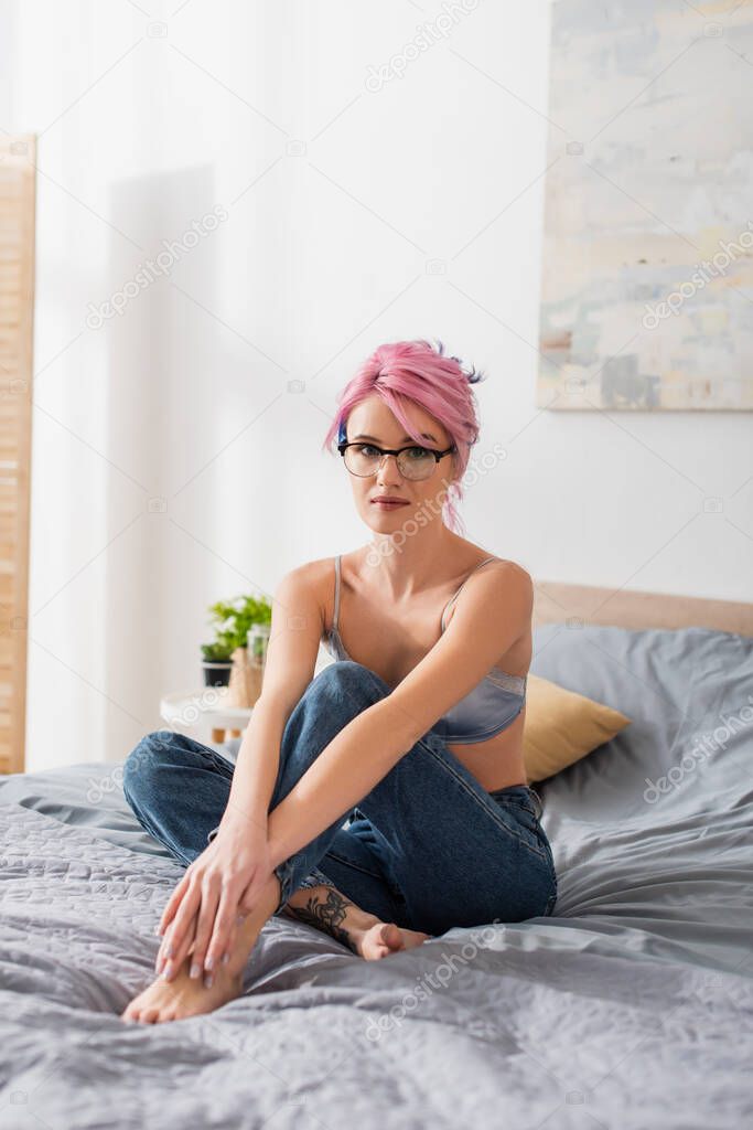 tattooed young woman with pink hair sitting in bra and jeans on bed 