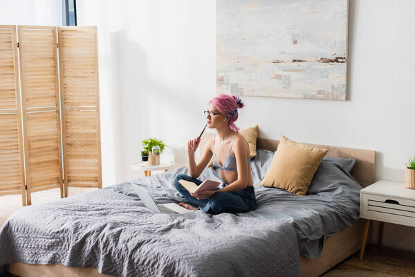 задумчивая молодая женщина с окрашенными волосами держит ноутбук во время учебы в Интернете дома