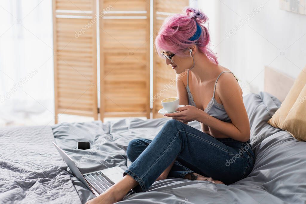 smiling young woman in earphone holding cup of coffee near gadgets on bed