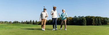 Kıdemli ırklar arası arkadaşlar golf sopalarıyla sahada yürüyor.