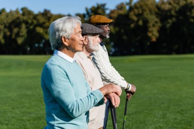 Golf sopalarıyla ırklar arası dostların yanında duran yassı şapkalı yaşlı adamın yan görüntüsü.