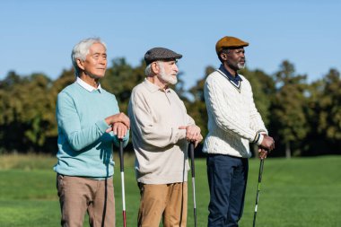 Irklar arası arkadaşlarının yanında golf sopalarıyla duran yassı şapkalı son sınıf öğrencisi.