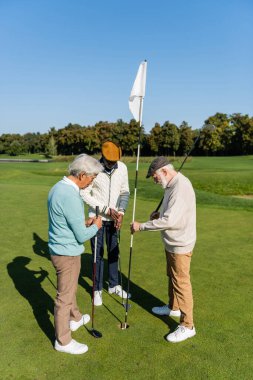 Düz şapkalı yaşlı adam golf sahasında çok ırklı arkadaşlarının yanında bayrak sallıyor. 
