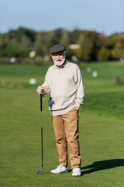 Düz şapkalı son sınıf öğrencisi cebinde golf sopasıyla yeşil alanda duruyor. 
