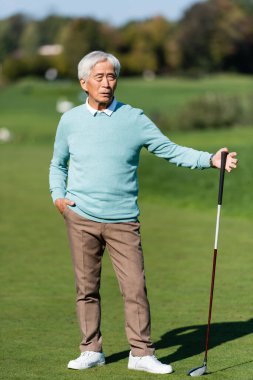 Asyalı son sınıf öğrencisi, yeşil alanda el ele ve golf sopasıyla ayakta duruyor.