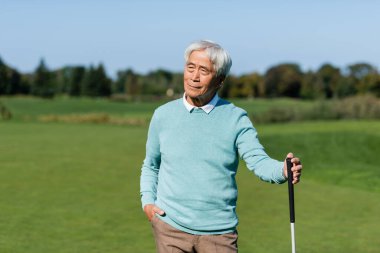 Asyalı son sınıf öğrencisi cebinde ve elinde golf sopasıyla yeşil alanda duruyor.