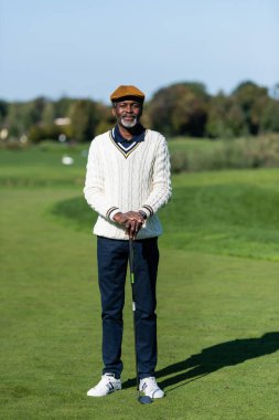 Orta yaşlı Afro-Amerikan bir adam elinde golf sopasıyla yeşil alanda duruyor.