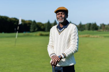 Neşeli Afrikalı Amerikalı adam golf sopasıyla yeşil sahada duruyor.