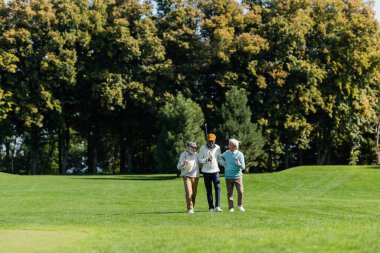 Yaşlı çok ırklı golfçüler ağaçların yanındaki yeşil alanda golf sopalarıyla yürüyorlar.
