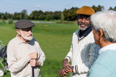Yaşlı çoklu etnik erkekler ellerinde golf sopaları ve bulanık Asyalı arkadaşlarıyla konuşuyorlar. 