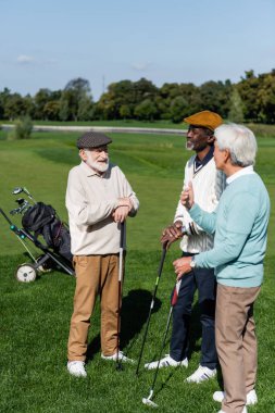 Kıdemli çoklu etnik arkadaşlar gülümsüyor ve yeşil çimlerde golf sopaları tutuyor
