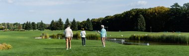 Golf sopalarıyla gölün kenarında yeşil çimlerde yürüyen yaşlı çoklu etnik erkeklerin arka görünüşü, afiş.