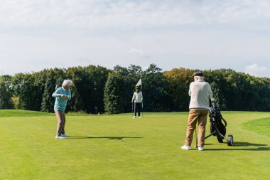 Kıdemli Asyalı adam bayrak sopası ve golf arabasıyla ırklar arası arkadaşlar arasında golf oynuyor.