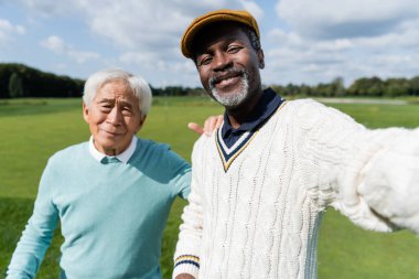 Gülümseyen Afrikalı Amerikalı adam golf sahasının kıdemli Asyalı arkadaşıyla selfie çekiyor. 