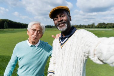 Memnun Afrikalı Amerikalı adam golf sahasının kıdemli Asyalı arkadaşıyla selfie çekiyor. 