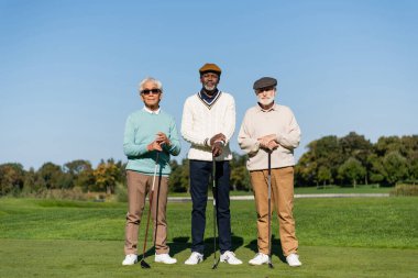Güneş gözlüklü üst düzey Asyalı adam golf sopalı çok ırklı arkadaşlarının yanında duruyor. 