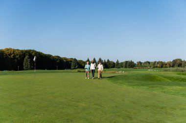 Güneş gözlüklü Asyalı adam golf sahasında çok ırklı arkadaşlarıyla yürüyor.