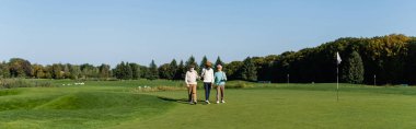 Güneş gözlüklü Asyalı adam golf sopaları ve pankartıyla çok ırklı üst düzey arkadaşlarının yanında yürüyor.