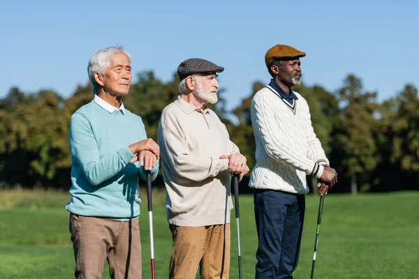 senior man in flat cap near interracial friends standing with golf clubs