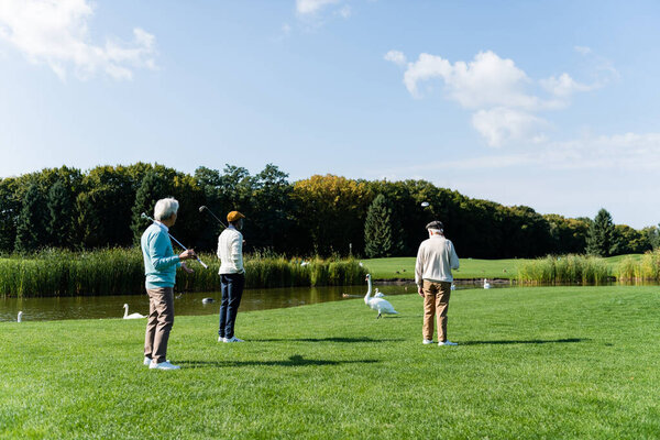 back view of senior multiethnic friends with golf clubs standing on green lawn near pond 