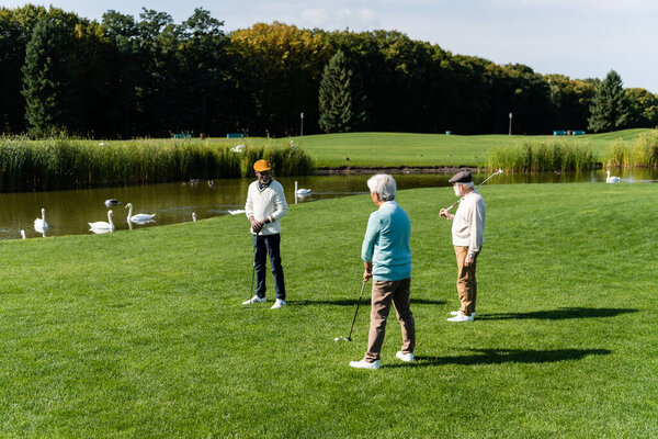 senior multiethnic men playing golf near pond with swans 