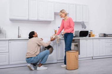 happy blonde woman passing dirty clothes to cheerful african american man clipart