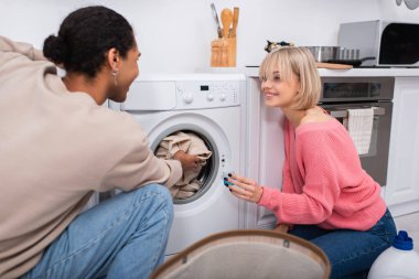 Afro-Amerikalı adamın çamaşır makinesine çamaşır koyduğu yerde elinde deterjanı olan neşeli sarışın kadın.