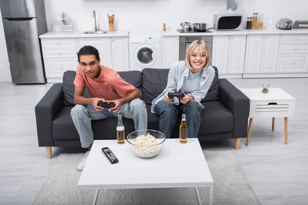 KYIV, UKRAINE - DECEMBER 6, 2021: cheerful multiethnic couple playing video game in living room 
