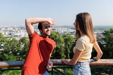 Happy man looking at girlfriend near railing on viewpoint in city  clipart