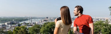Positive tourist looking away near brunette girlfriend with binoculars on viewpoint in city, banner  clipart