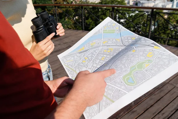Cropped view of tourist pointing at map near girlfriend with binoculars outdoors