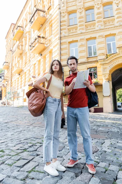 stock image Traveler pointing at map near girlfriend with backpack and binoculars on urban street 