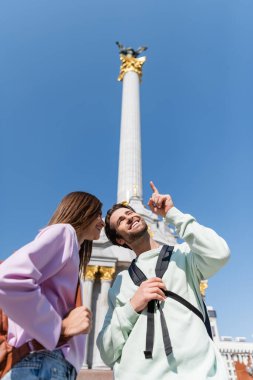 KYIV, UKRAINE - 1 Eylül 2021: Independence Meydanı 'nda kız arkadaşını işaret eden sırt çantalı gülen gezginin düşük açılı görüntüsü
