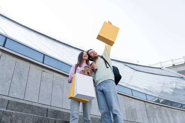 Low angle view of happy travelers with coffee to go, backpacks and shopping bags standing on urban street