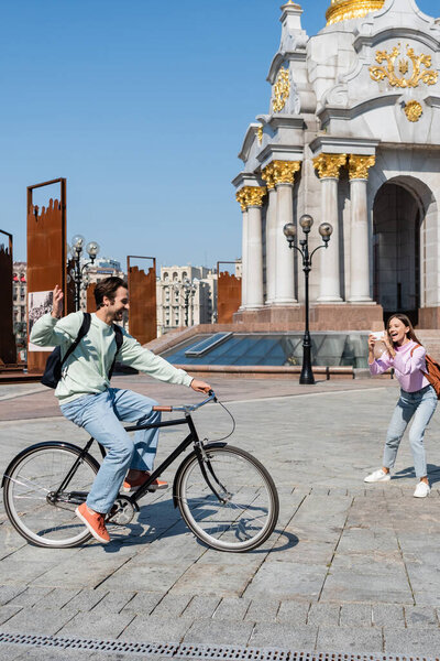 KYIV, UKRAINE - SEPTEMBER 1, 2021: Cheerful woman taking photo on smartphone while boyfriend riding bike on urban street, Independence Square