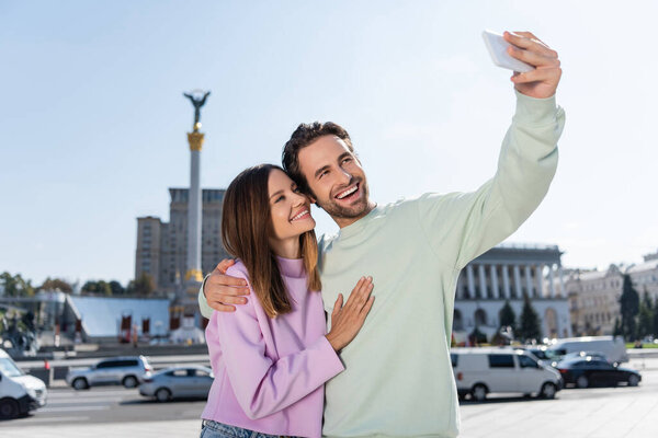 KYIV, UKRAINE - SEPTEMBER 1, 2021: Cheerful couple hugging and taking selfie on smartphone on Independence Square