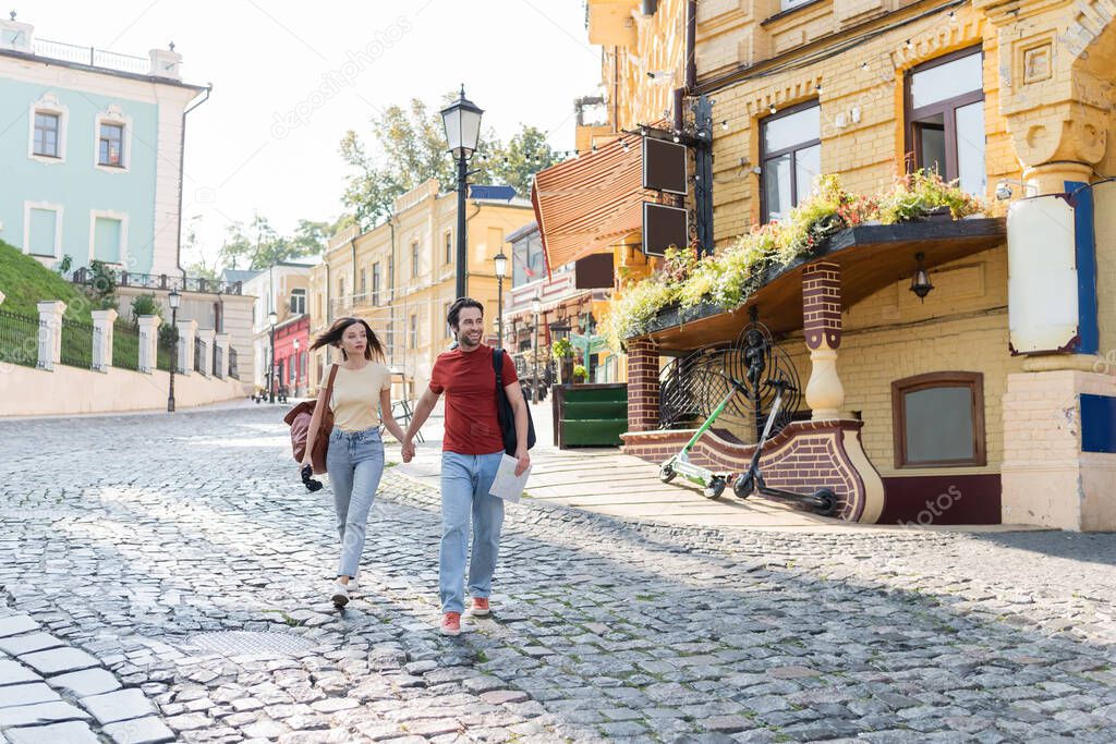 Happy tourist holding map and hand of girlfriend while walking on urban street 