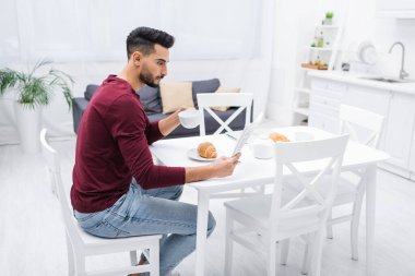 Side view of arabian man reading news and holding cup near breakfast in kitchen 