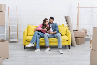 Positive interracial couple holding bengal cat on couch near carton boxes at home 