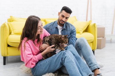 Positive woman holding bengal cat near arabian boyfriend at home 