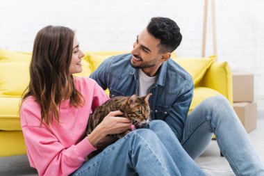 Positive muslim man looking at girlfriend with bengal cat at home 