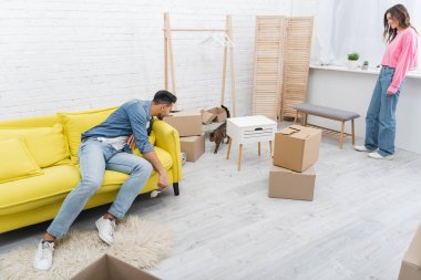 Young interracial couple looking at bengal cat near packages in living room 